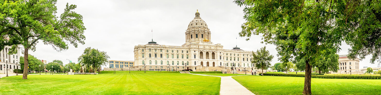 Front view of Minnesota Capitol building on a bright day with green grass, leafy trees, and visitors climbing the front steps.