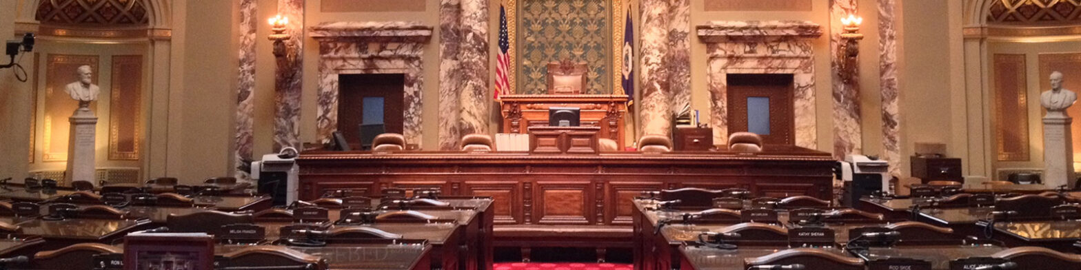 Empty Minnesota Capitol chamber with empty seats, the American and Minnesota state flags, and statues of prominent former Minnesota legislators.